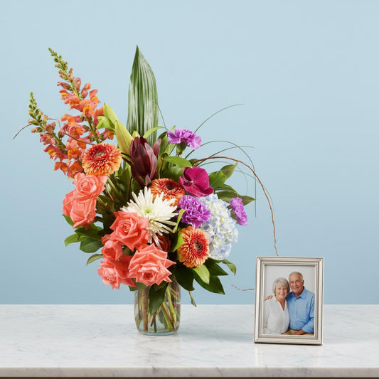 Colorful flower arrangement in a vase on a table with a framed photo of an elderly couple.