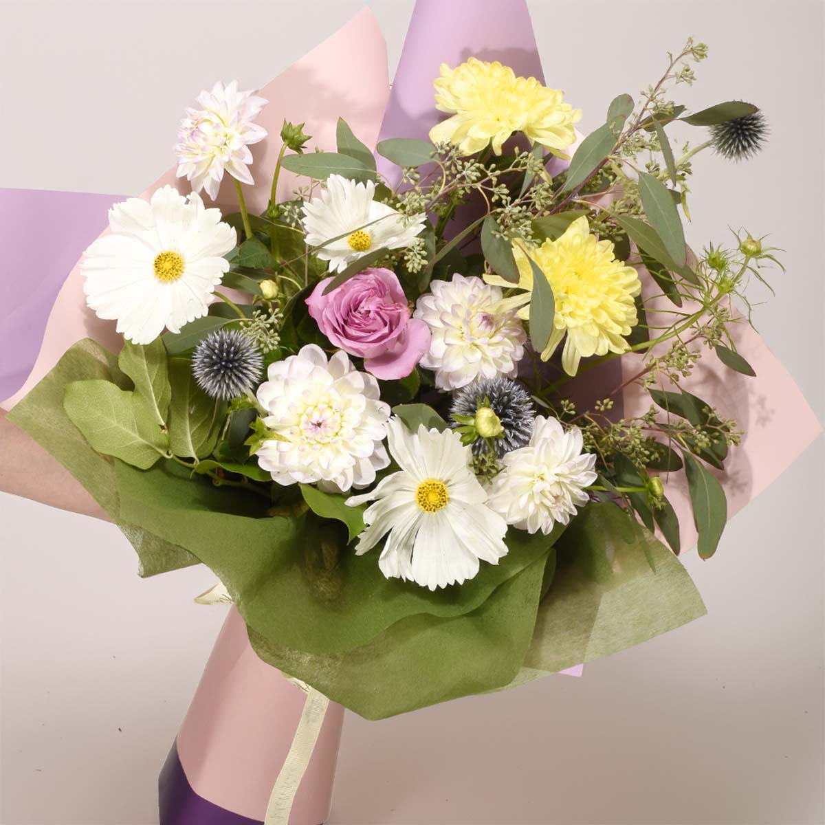A vibrant floral bouquet featuring white daisies, yellow chrysanthemums, a pink rose, and white dahlias, wrapped in green and pink tissue paper with a pink cylindrical vase or holder visible at the base.