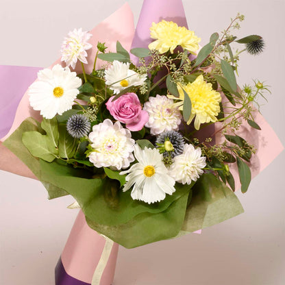 A vibrant floral bouquet featuring white daisies, yellow chrysanthemums, a pink rose, and white dahlias, wrapped in green and pink tissue paper with a pink cylindrical vase or holder visible at the base.